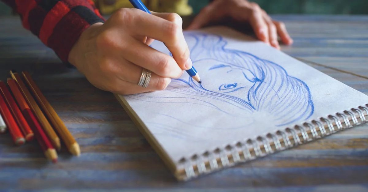 Gros plan d'une main esquissant un portrait sur un carnet de croquis spirale avec des crayons de couleur sur une table.