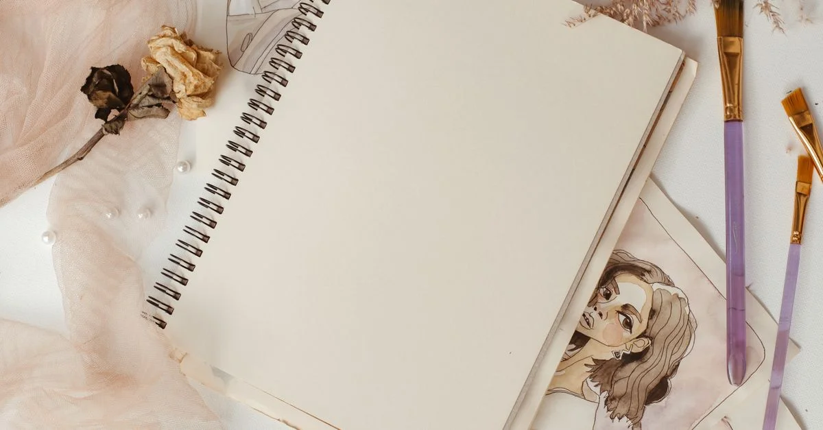 Composition élégante de fournitures artistiques incluant un carnet de croquis, des pinceaux, et des fleurs séchées sur une surface blanche.